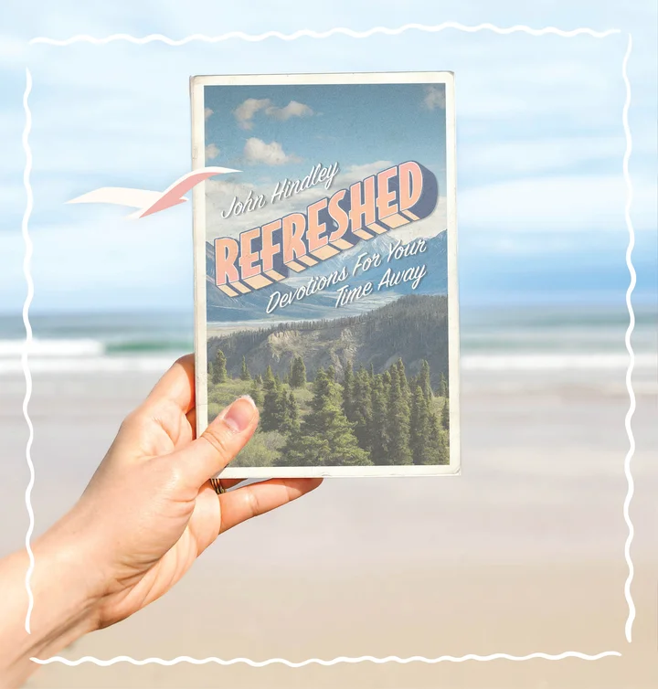 Photograph featuring a person holding up Refreshed: Devotions for Your Time Away with a beach scene in the background